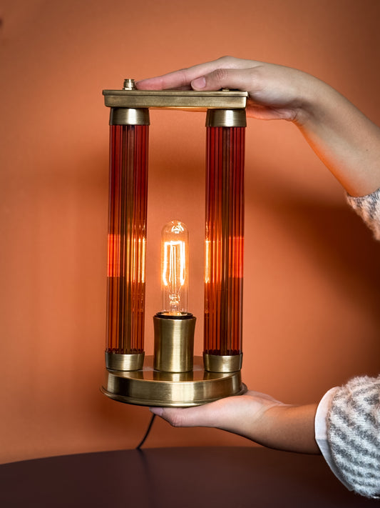 A hand holding a table lamp with a brass structure and gradient glass, featuring an edison-style bulb illuminated.